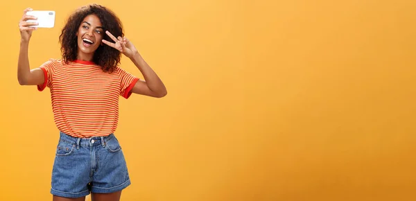 Girl making video vlog with brand new smartphone posting in internet trying become famous standing over orange background posing for selfie gazing at gadget screen showing peace or victory gesture
