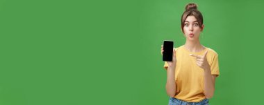 Meraklı kadın portresi bekleyemez. Yeni akıllı telefonu kullan ve cep telefonu ekranını işaret et. Dudakları heyecan ve ilgi ile kıvır ve yeşil duvardaki yeni cihaz modelini tanıt.