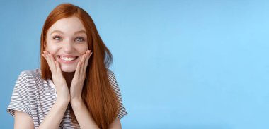 Heyecanlı, mutlu, kızıl saçlı, neşeli, mutlu, yanaklarına dokunarak gülümseyen kız şaşırtıcı bir şekilde muhteşem bir doğum günü hediyesi aldı. Şaşırtıcı bir zafer, sevindirici, mavi arka plan. Boşluğu kopyala