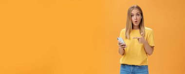 Akıllı telefonlara bağlanan kablosuz kulaklıklardan etkilenen adam ekranı işaret ederek sorgulanıyor ve şaşırtılan kıvrılmış dudaklar turuncu duvarın üzerinde kulaklık takan kameraya bakıyor.