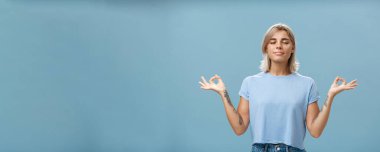 Meditasyon sırasında içsel benliği dinlemek. Sakin, mutlu ve rahat, bronzlaşmış, kollarında dövmeleri olan sarışın bir kadın. Mavi duvarın üzerinde duruyor. Zen hareketleriyle yoga yapıyor..