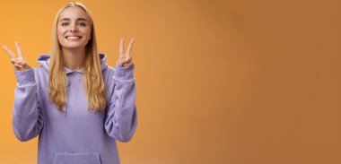 Yaşam tarzı. Barışçıl, mutlu, çekici, sarışın Avrupalı, kapüşonlu, gülümseyen, barışçıl zafer hareketi yapan genç kız öğrenci. Sevgi ve mutluluk, ayakta duran hippi işareti turuncu arka plan..