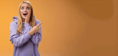 Şaşırtıcı derecede heyecanlı kapüşonlu genç sarışın kız avucunun içi gibi bağırıyor sağa doğru hayret verici bir şekilde haykırıyor ünlü süper star neşeli, pozitif duyguları tutamıyor, turuncu arka plan.