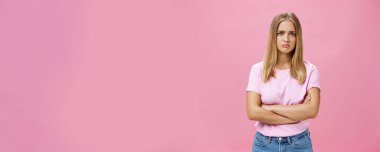 Üzücü kıskançlık ve hayal kırıklığına uğramış kasvetli, açık renk tişörtlü, kollarını göğsüne dolamış, dudaklarını büzmüş, endişeli ve pembe duvardan dolayı üzgün görünen genç bir kadın.