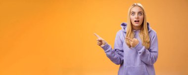 Kapüşonlu, sarışın, şaşkın bakışlı, şaşırmış, şaşırmış bakışlı, ağzı açık, solu işaret eden, dedikoducu, turuncu arka planda duran sarışın kadın şaşırmış..