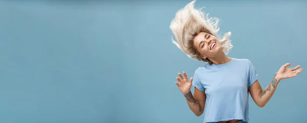 Waist-up shot of artistic and creative attractive carefree blonde female waving head and hair joyfully dancing and jumping having fun smiling broadly over blue background. Emotions and people concept
