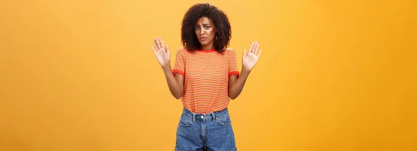 Silly insecure and sad dark-skinned female model in trendy striped t-shirt and shorts raising arms in surrender frowning being uninvolved and unaware standing clueless over orange background