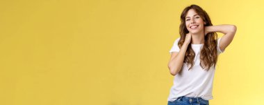 Neşeli, karizmatik, güzel, kıvırcık saçlı, esnemiş, sarı arka planı olan, boynuna dokunmuş, sırıtan, gülümseyen, rahatlatıcı, yaz tatili rezervasyonu yapan bir kadın.