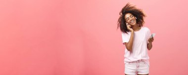 Kulağındaki güzel melodilerle endişelenmekten kurtuluyorsun. Moda kıyafetleri içinde, kulaklıkla müzik dinleyen, kulaklıklara dokunan, gülümseyen, dans eden ve elinde akıllı telefon tutan çekici bir Afrikalı kadın.