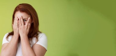 Dikizlemiyorum. Karizmatik orta yaşlı kızıl saçlı kadın avuçlarının içine bak parmaklarının arasından bak bir göz kamerasının ilgisini çek sürpriz bir hediye bekle yeşil arka plan.