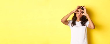 Mutlu ve komik Afro-Amerikan kızı tasviri, parmak gözlüğü yapıyor ve onlara bakıyor, sarı arka planda duran inanılmaz bir şeye..