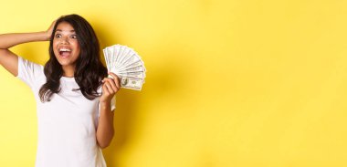 Şanslı Afro-Amerikalı kız resmi, sol üst köşede heyecanlı görünüyor, para tutuyor, alışverişe gidiyor, sarı arka planda duruyor..