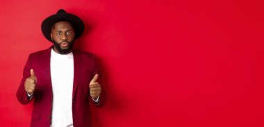 Etkilenmiş siyahi adam başparmağını kaldırıyor ve kameraya bakıyor, onaylıyor ve övüyor güzel, kırmızı arka plan.