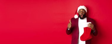 Etkilenmiş Afrikalı Amerikalı adam elinde hediyelerle Noel çorabı tutuyor, başparmağını kaldırıyor, şaşırmış görünüyor, kırmızı arka planda duruyor..