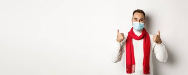 Covid-19, tecrit ve karantina konsepti. Yüz maskesi takan gülümseyen ve baş parmağını kaldıran, kendini koronavirüsten koruyan, beyaz arka planda duran.