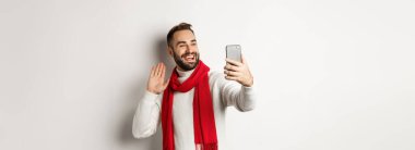 Yakışıklı sakallı adam arkadaşlarını arıyor, merhaba diyor ve cep telefonuna el sallıyor, süveter ve kırmızı eşarpla beyaz arka planda duruyor..