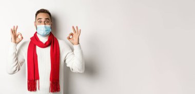 Covid-19, tecrit ve karantina konsepti. Yüz maskeli etkileyici bir adam, tamam işaretleri gösteriyor ve katılıyor, harika bir ürün öneriyor, beyaz arka plan..