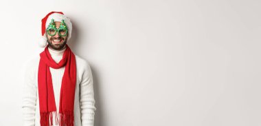 Noel, yeni yıl ve kutlama konsepti. Mutlu adam gülüyor, Noel Baba şapkası ve parti gözlüğü takıyor, beyaz arka planda duruyor..