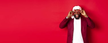 Komik siyah adam yeni yılı kutluyor, parti gözlüğü takıyor ve Noel Baba şapkası takıyor, kırmızı arka planda eğleniyor..