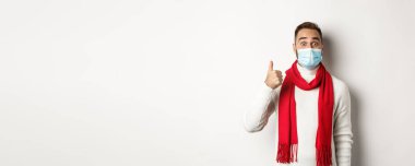 Covid-19, tecrit ve karantina konsepti. Tıbbi maskeli, şaşırtıcı ve tatmin olmuş bir adam başparmağını kaldırmış, heyecanlı görünüyor, iyi, beyaz bir arka plan gibi..