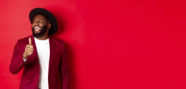 Yakışıklı Afro-Amerikan erkeği eğleniyor, başparmağını kaldırıyor ve iyi bir şakadan gülüyor, kırmızı arka planda duruyor..