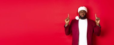 Noel, parti ve tatil konsepti. Sefil ve üzgün Afro-Amerikan adam parmaklarını havaya kaldırmış, hayal kırıklığına uğramış görünüyor, Noel Baba şapkası takıyor, kırmızı arka plan.