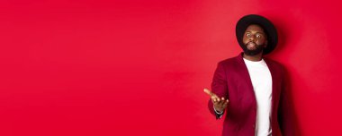 Şok olmuş Afro-Amerikan bir adam kameraya bakıyor ve üzgün bir şeye işaret ediyor, parti ceketi ve şapkası içinde duruyor, kırmızı arka plan.