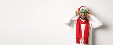 Noel, yeni yıl ve kutlama konsepti. Mutlu adam gülüyor, Noel Baba şapkası ve parti gözlüğü takıyor, beyaz arka planda duruyor..