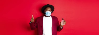 Covid-19, karantina ve tatil konsepti. Tıbbi maskeli, el dezenfektanı ve baş parmağını kaldırmış, antiseptik kullanmayı öneren Afrikalı genç bir Amerikalı..