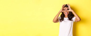 Mutlu ve komik Afro-Amerikan kızı tasviri, parmak gözlüğü yapıyor ve onlara bakıyor, sarı arka planda duran inanılmaz bir şeye..