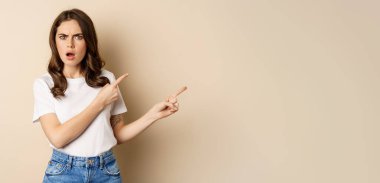 Frustrated woman looking insulted, pointing fingers right and gasping shocked, standing in t-shirt over beige background.
