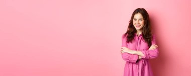 Bahar elbisesi giymiş güzel bir kadın, göğsünde el ele tutuşmuş ve kendine güvenen bir ifadeyle gülümsüyor, pembe arka planda mutlu duruyor..