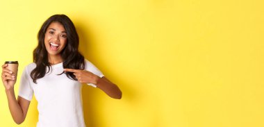 Afro-Amerikalı neşeli bir kadın tasviri, kafeden iyi kahve öneriyor, fincanı işaret ediyor, sarı arka planda duruyor..