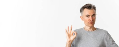 Gri kazaklı, orta yaşlı, tatmin olmuş bir adam. Onaylama işareti gösteriyor. İyi bir şeyi övüp beyaz arka planda duruyor..