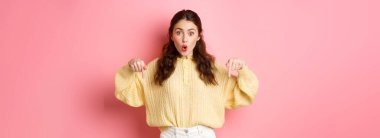 Vay canına, süper havalı. Heyecanlı esmer kız parmaklarını aşağı işaret ediyor, nefes nefese merak ediyor, yön gösteriyor, alt logoda işaret ediyor, pembe arka planda duruyor..