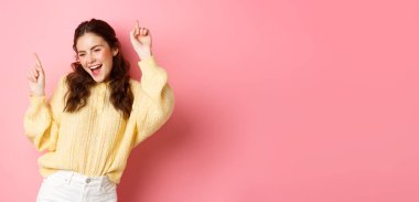 Mutlu, çekici bir kadın eğleniyor, parmaklarını havaya kaldırıyor, gamsızca dans ediyor, neşeli bir şekilde gülümsüyor, pembe arka planda duruyor..