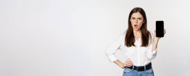 Shocked and frustrated woman showing mobile phone screen app, smartphone application, looking insulted, standing over white background.