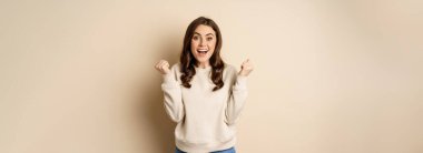 Enthusiastic brunette woman laughing and smiling, cheering, shaking hands and celebrating, triumphing, standing over beige background.