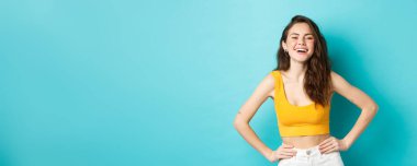 Kendine güvenen ve mutlu stil sahibi bir kadının gülüşü, el ele tutuşması ve gülümsemesi, yaz kıyafetleri içinde tasasız durması, mavi arka plan..