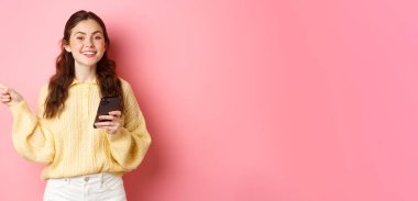 Güzel, modern bir kız akıllı telefonu elinde tutuyor ve bir kenara işaret ediyor. Cep telefonu kullanırken tanıtım metnini gösteriyor. Pembe arka planda duruyor..