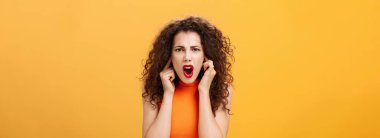 İncinmiş, gergin ve kendinden emin olmayan bir kadının belden yukarısından. Turuncu başlıklı kıvırcık saçlı, kapalı kulaklı parmakları kaşlarını çatmış turuncu duvarın üstünde gürültülü bir yerde dikilirken aynı soruyu tekrar soran..