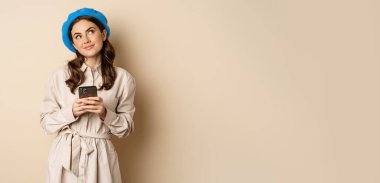 Dreamy smiling woman in stylish trenchcoat, looking up fantasizing while shopping on mobile phone app, using smartphone and thinking, standing over beige background.