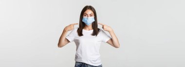 Covid-19, sağlık ve sosyal uzaklık kavramı. Tıbbi maskeli çekici esmer kız, beyaz arka planda, yüzü işaret ediyor..