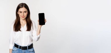 Confident woman in corporate clothes, showing smartphone screen, mobile interface of an application, standing over white background.