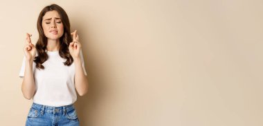 Hopeful woman praying, cross fingers for good luck, making wish, standing over beige background.