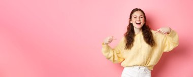 Kendinden emin mutlu kız kendini gösteriyor ve gülüyor, kendini tanıtıyor ve gülümsüyor, pembe arka plana karşı duruyor..