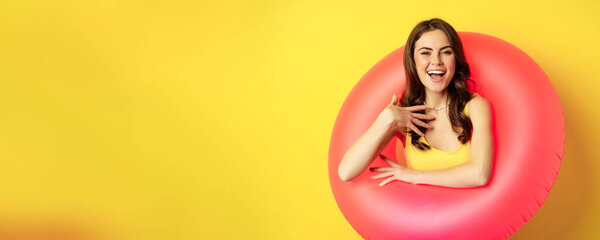 Close up portrait of beautiful brunette woman model inside pink swimming ring, beach accessories and summer vacation concept, yellow background.