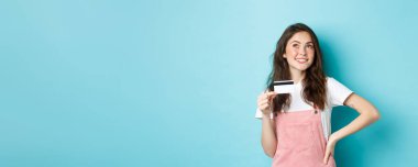 Alışveriş yapmayı düşünen düşünceli bir kız, elinde plastik bir kredi kartı tutarken düşünceli bir yüz ifadesiyle, ne alacağına karar verirken, mavi arka planda dururken..