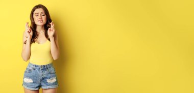Hopeful young beautiful woman praying, believe, hoping to receive smth, cross fingers for good luck, anticipating, standing over yellow background.