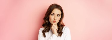 Close up portrait of coquettish smiling woman, glamour girl thinking, looking thoughtful, standing over pink background.